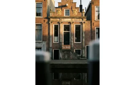 Historisch Amsterdams grachtenpand met sierlijke trapgevel en te koop-bord op de stoep, weerspiegeld in donker kanalwater.