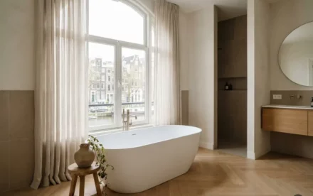 Vrijstaand wit bad onder groot raam in lichte Amsterdamse badkamer met eiken vloer, keramische vaas en hangende plant.