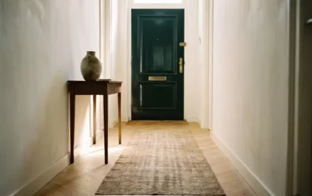 Zonnige Nederlandse hal met lichte eiken visgraatparket, witte muren, keramische vaas op sidetable en voordeur met messing beslag.
